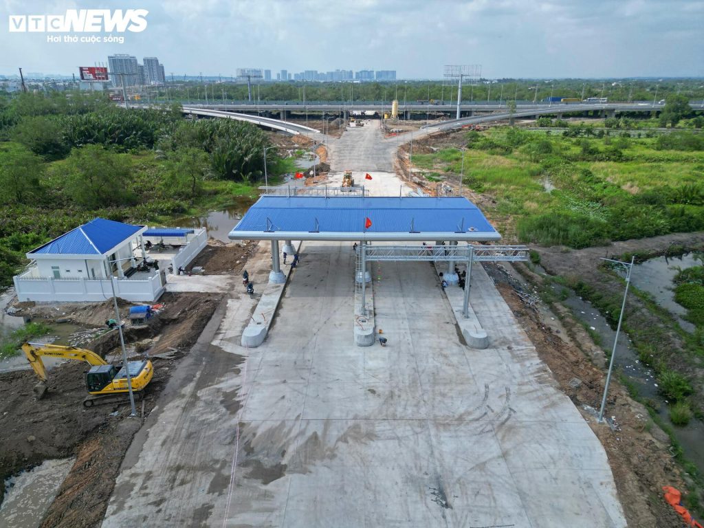 Tại khu vực nút giao Vành đai 3 với cao tốc TP.HCM - Long Thành - Dầu Giây, phần lớn bề mặt đường đã được thảm bê tông nhựa, các làn xe và vạch phân cách rõ ràng. Hệ thống trạm thu phí với mái che, khung biển báo gần như hoàn thiện, chỉ còn một số hạng mục phụ trợ đang được thi công.